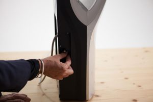 La mano di una persona collega un cavo bianco al lato di un moderno asciugamano elettrico Dualflow Plus nero e argento, appoggiato su un tavolo di legno chiaro.