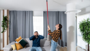 Una donna usa uno straccio per raggiungere il soffitto, mentre un uomo è seduto su un divano e si tappa le orecchie, con aria frustrata. Nelle vicinanze, un elegante dispenser di sapone e asciugamani piegati con cura aggiungono stile al moderno arredamento del loro soggiorno.
