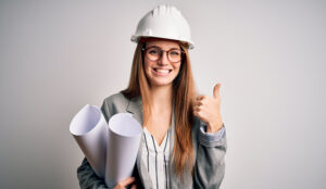 Una donna sorridente che indossa un casco da costruzione bianco e degli occhiali tiene in una mano dei progetti architettonici e con l'altra alza il pollice, in piedi su uno sfondo semplice, pronta a progettare spazi moderni con le soluzioni Asciugamani Elettrico.