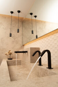 Bagno moderno con lavandino in pietra beige, rubinetti neri, lampade a sospensione nere, parete con piastrelle esagonali geometriche, grande specchio e una scultura decorativa dorata sul bancone. Un asciugamano elettrico è installato nelle vicinanze per un'asciugatura rapida delle mani.