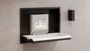 Fasciatoio a parete con piattaforma bianca ribaltabile e dispenser di sapone grigio sopra, decorato con illustrazioni di palloncini colorati e istruzioni di sicurezza. Il fasciatoio è posizionato contro una parete di colore chiaro.