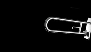 Una barra di sicurezza in metallo è montata su una parete su uno sfondo nero. La barra ovale e lucida è posizionata vicino a un dispenser di asciugamani, con una staffa di supporto fissata alla parete sul lato destro.