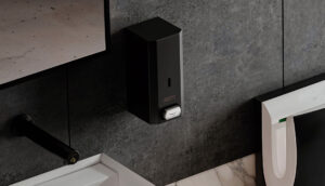 A dispenser di sapone nero è montato su una parete di piastrelle grigio scuro in bagno, tra uno specchio e un orinatoio, con parte di un lavandino e rubinetto visibili sotto.
