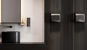 Bagno moderno con lavandino nero, uno specchio sopra, un piccolo vaso con bastoncini di canna, un dispenser di sapone a parete e due dispenser di carta mani neri su una parete scura e strutturata.