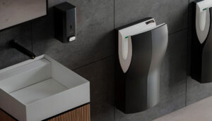 Moderno bagno pubblico con lavandino rettangolare bianco, dispenser di sapone nero e asciugamani ad alta velocità montati su una parete piastrellata grigia. Sulla destra è visibile parte di un orinatoio.
