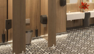 Moderno bagno pubblico con cabine in legno, pavimento piastrellato con motivi floreali bianchi e neri, un lavandino bianco con un dispenser di carta mani, un cestino per i rifiuti e un vaso di fiori rossi sul bancone.