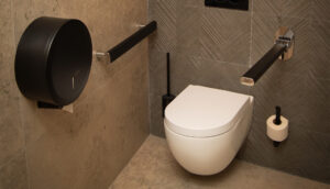 Bagno accessibile con WC sospeso, due maniglioni orizzontali, un grande distributore di carta igienica nera, un distributore di carta mani per maggiore comodità, uno scopino per WC, pareti e pavimento piastrellati chiari.