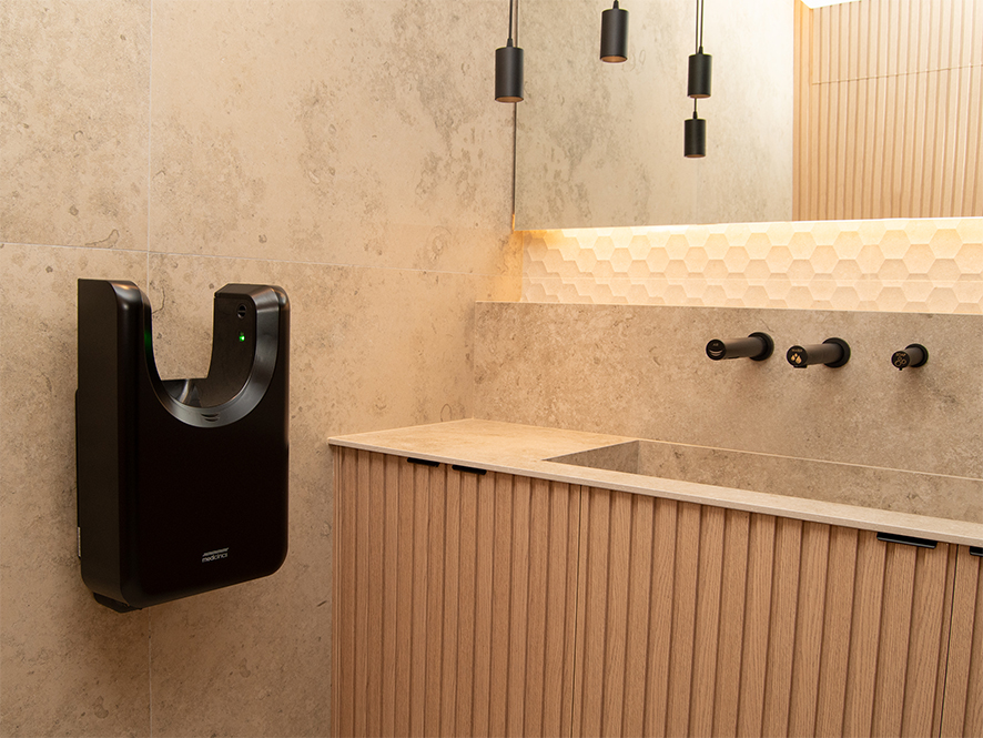 Un bagno moderno con un asciugamani nero montato a parete, un lavandino rettangolare con tre rubinetti neri e pannelli in legno chiaro. Un grande specchio e un'illuminazione soffusa creano un look pulito e minimalista.