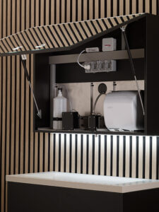 Un moderno mobiletto a parete, "Armadietto con 3 moduli: erogatore di sapone + erogatore d'acqua + asciugamani (rif. BTM 02)", è appoggiato su doghe verticali in legno sopra un elegante piano di lavoro bianco.