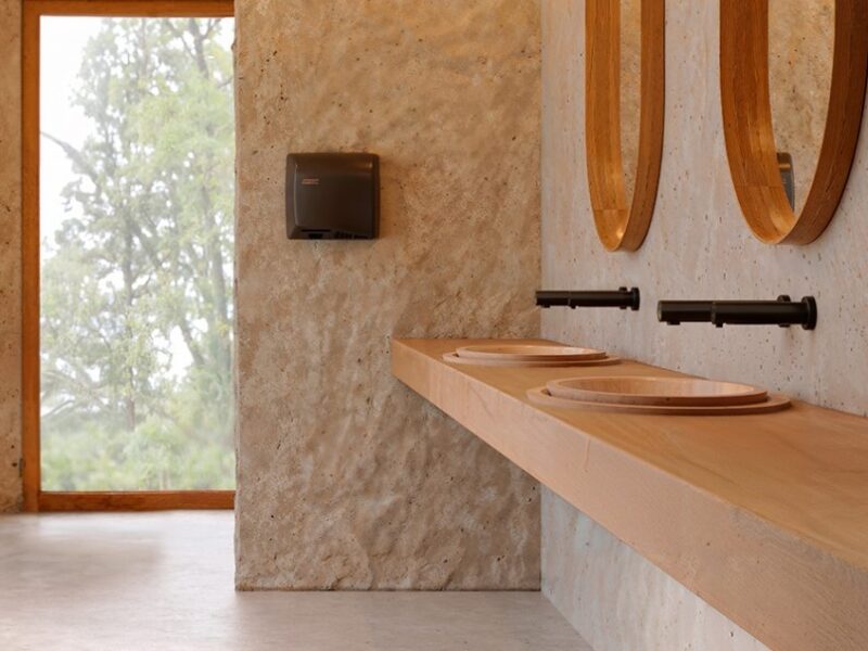 Bagno moderno con pareti in pietra testurizzata, piani di lavoro in legno con due lavandini rotondi, rubinetti neri, specchi ovali e un'ampia finestra che mostra il verde esterno. Sulla parete è montato un distributore di asciugamani di carta.