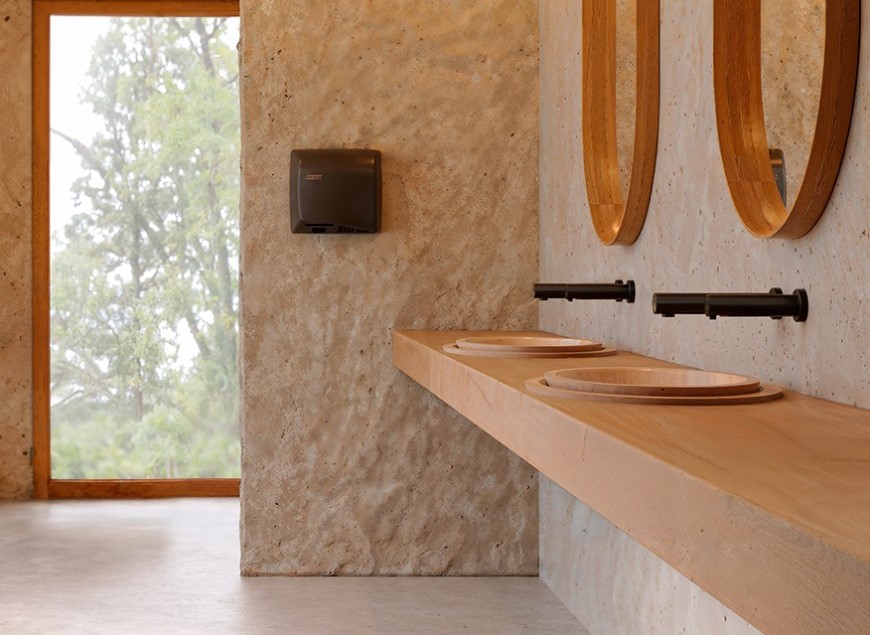 Bagno moderno con pareti in pietra testurizzata, piani di lavoro in legno con due lavandini rotondi, rubinetti neri, specchi ovali e un'ampia finestra che mostra il verde esterno. Sulla parete è montato un distributore di asciugamani di carta.