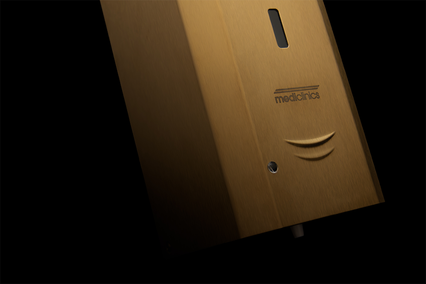 Primo piano di un dispenser Mediallnics montato a parete, di colore oro, con un pulsante rettangolare e una piccola finestra, su sfondo nero.