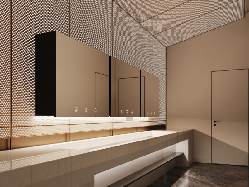 Un bagno moderno e minimalista con pareti beige, un lungo piano di lavoro rettangolare, grandi specchi e illuminazione sotto il mobile. La stanza presenta un soffitto in metallo perforato e una semplice porta chiusa sulla destra.