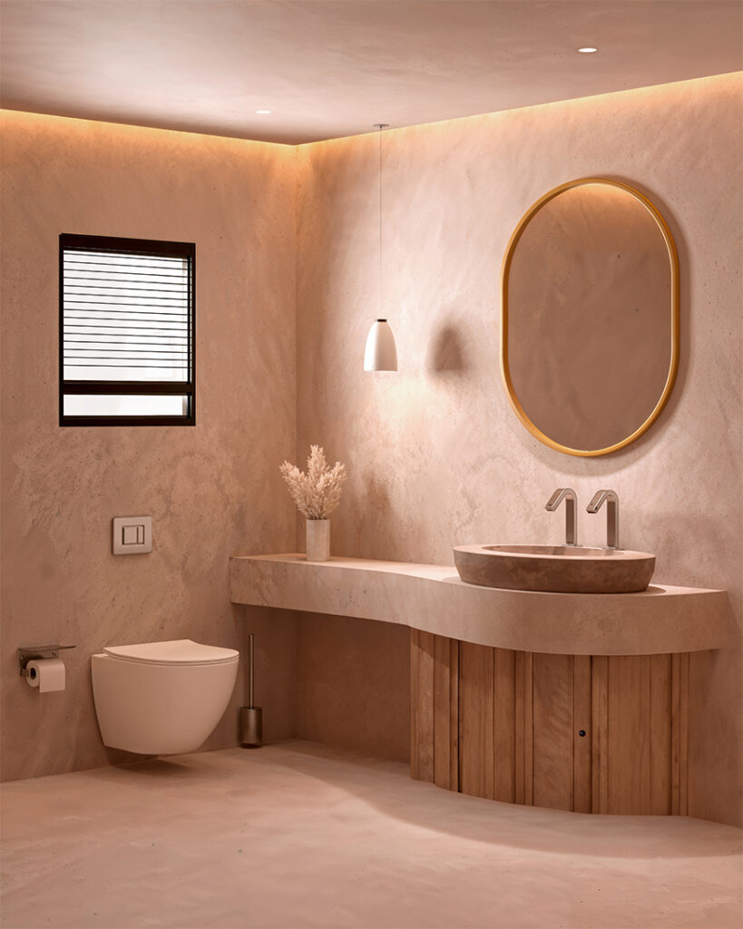 Bagno minimalista con pareti beige tenui, wc galleggiante, mobiletto in legno con specchio ovale, lavandino a vaschetta, luce a sospensione, finestra con persiane e vaso con fiori secchi. L'illuminazione soffusa a incasso aggiunge calore.
