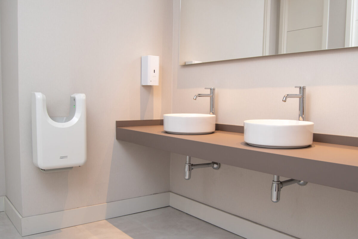 Un bagno moderno con due lavandini bianchi rotondi su un piano d'appoggio beige, rubinetti, un grande specchio a parete e un asciugamani a muro. Il design è minimalista e pulito.